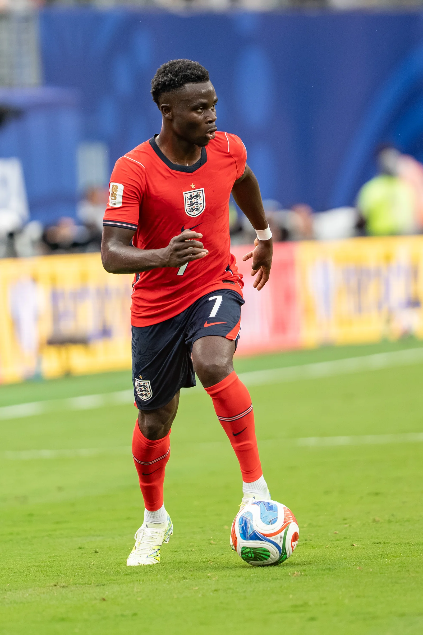 Bukayo Saka in a red England kit with the ball on a football pitch.
