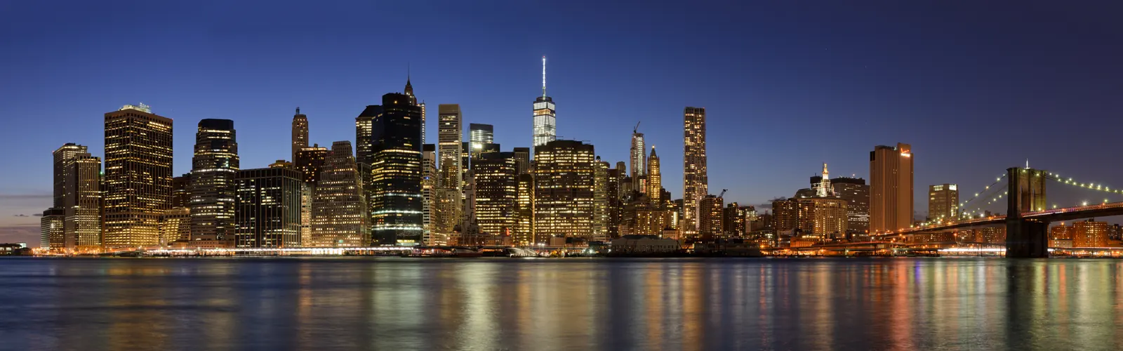 Lower Manhattan and the Brooklyn Bridge illuminated across the East River at dusk.